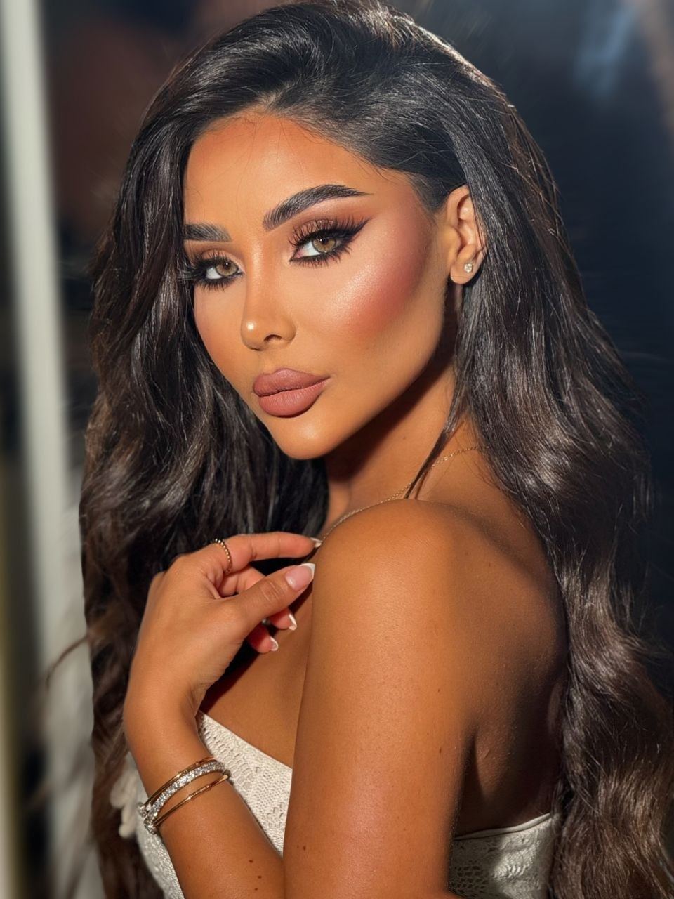 Zina Almokri in a warm, softly lit beauty portrait, looking at the camera with her hand resting on her shoulder